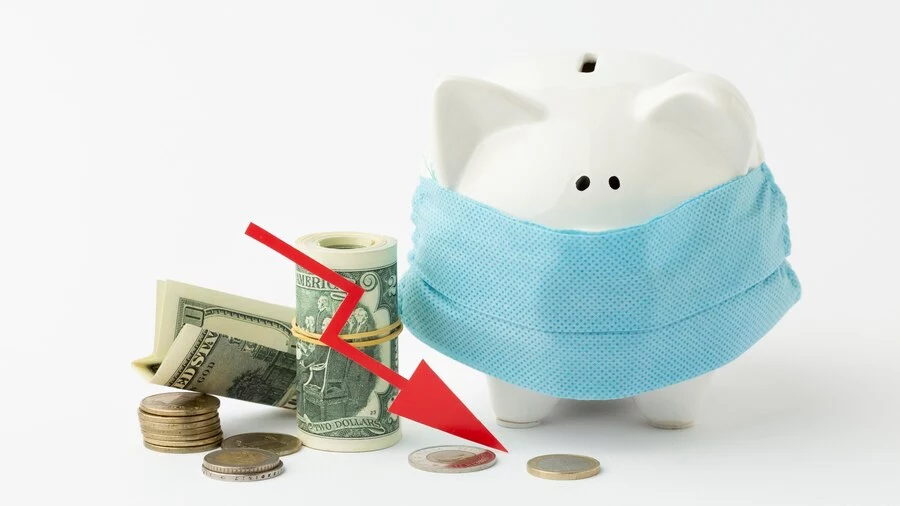 A white piggy bank wearing a blue surgical mask stands on a white surface. Next to it, a red downward-pointing arrow pierces through a rolled stack of dollar bills. Scattered coins and additional dollar bills are also visible. The image symbolizes economic downturn, financial health concerns, or financial loss, particularly in the context of a crisis.