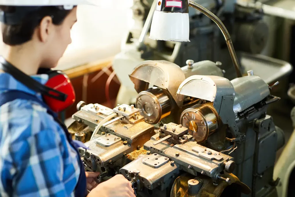 A person wearing a white hard hat, ear protection, and a blue plaid shirt is intently working on complex industrial machinery. Their hands are on the controls, and metal shavings are visible on the machine bed, indicating a manufacturing or workshop environment. The scene highlights skilled labor and industrial processes.